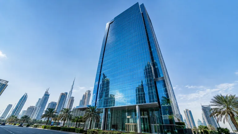 A glass-fronted bank office tower in Dubai reflecting the blue sky, seen from street level